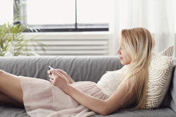 A picture of a beautiful young blonde woman holding a digital tablet and lying on the couch - Powered by Adobe