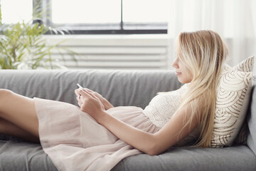 A picture of a beautiful young blonde woman holding a digital tablet and lying on the couch - Powered by Adobe