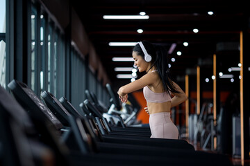 Fototapeta na wymiar Side view of beautiful young asian woman running on treadmill and listening to music via headphone with arm a smart watch for tracking speed during sports training in a gym.