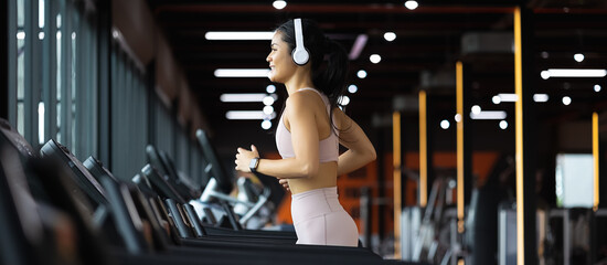 Panoramic Side view of beautiful young asian woman running on treadmill and listening to music via headphone with arm a smart watch for tracking speed during sports training in a gym.