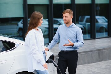 Happy young adult man and smiling woman charging electric car.