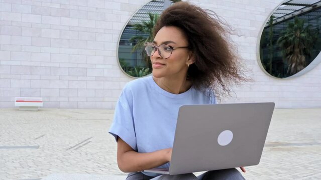 Beautiful positive confident African American curly haired young woman, with glasses, manager, company ceo, freelancer, sits with laptop outdoors near business center, work online, smiles friendly