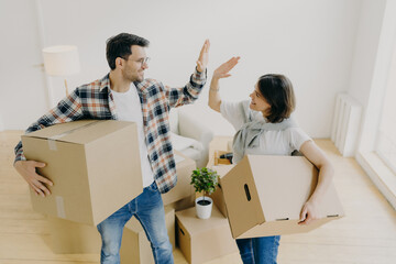 Photo of lovely husband and wife express their agreement, carry cardboard boxes with personal belongings, move in new apartment, pose in empty messy room, decide about interior of living room