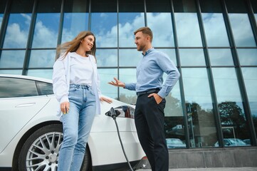 Fototapeta premium Young couple man and woman traveling together by new car having stop at charging station.