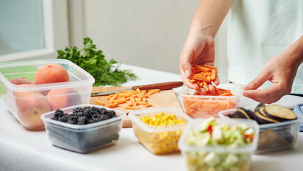 Woman holding plastic container with slices of carrot at table in kitchen. Raw vegetables for freezing for winter storage in trays.