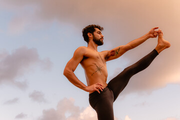 Young beautiful man practicing ashtanga yoga at sunset on the beach. Doing exercises and stretching. Extended Hand to Big Toe Pose