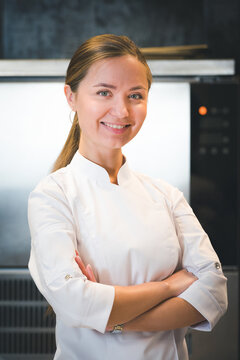 Portrait of confident and smiling young woman chef dressed in white uniform, professional kitchen are on background. Restaurant  kitchen