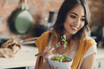 A young, beautiful, charming woman eats fresh vegetable salad in the kitchen at home