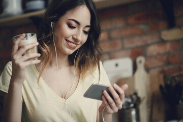 Young, charming, brunette girl watching the movie on her phone in headphones and holding a cup of...