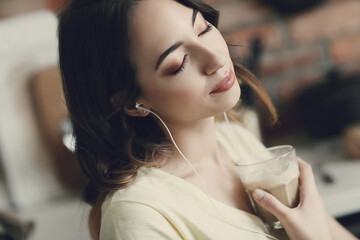 Young, charming, dark-haired girl listening to music in headphones and holding a cup of coffee - obrazy, fototapety, plakaty