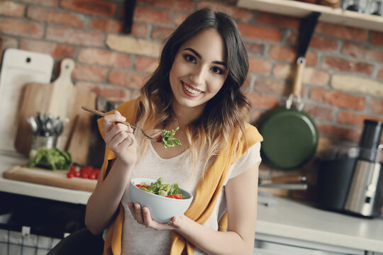 A young, beautiful, charming woman with fresh vegetable salad looking into the camera at the kitchen at home