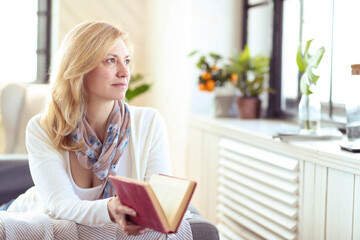 A blonde middle-aged woman holding a book and looking out the window