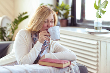 A blonde middle-aged woman sitting in a couch and drinking hot,delicious cup of tea
