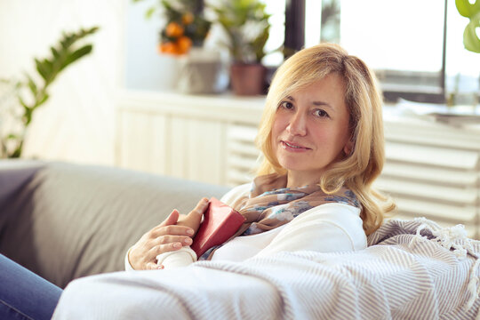 A blonde middle-aged woman sitting in a couch and holding her favorite book at home