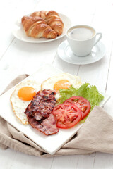 Morning breakfast with fried eggs,bacon,sliced vegetables,croissants and a cup of coffee cappucino on a wooden background