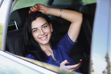 Photo of a young, brunette woman in a blue dress holding mobile phone and posing in her car