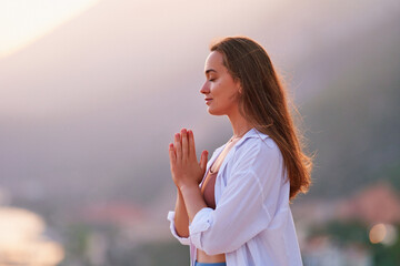 Inspired hopeful serene female meditate with closed eyes and standing alone with praying hands at sunset. Relief stress and clean healthy minds