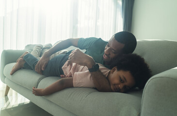 Family spending time at home. Happy African American father and little boy cuddling lying on couch with evening sunbeams shine down in living room at home. Sweet moments of fatherhood concept.