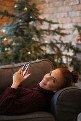 Cute, pretty girl with a bun on her head in a cozy sweater lying on the couch with a phone