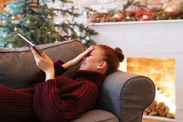 Cute, pretty girl with a bun on her head in a cozy sweater lying on the couch with a digital tablet