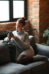 A cute, pretty girl with a bun on her head in a cozy sweater sitting on the couch with a digital...