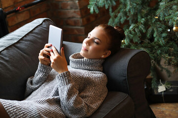 A cute, pretty girl with a bun on her head in a cozy sweater lying on the couch with a digital...