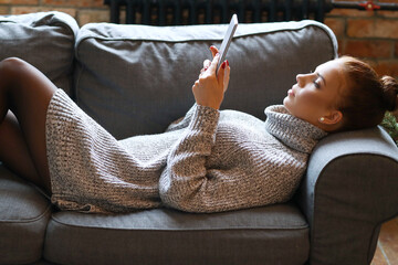 A cute, pretty girl with a bun on her head in a cozy sweater lying with a digital tablet on the sofa