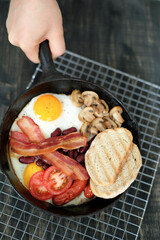 Top view of man`s hand holding pan of English breakfast with fried eggs, bacon, mushrooms,bread and vegetables .