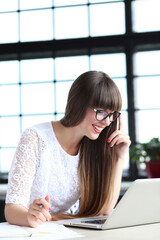 Close-up photo of a young,attractive girl in glasses with bangs working on the computer and talking on the cell phone against windowsill.