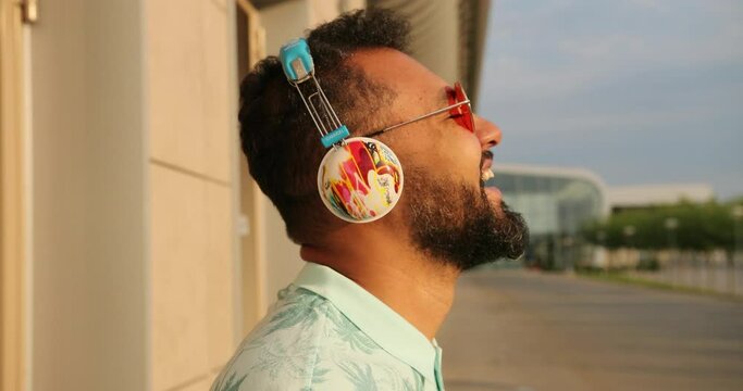 Relaxed afro-american man in stylish pink sunglasses and headphones is laughing near the airport terminal. 4k footage. Side portrait.