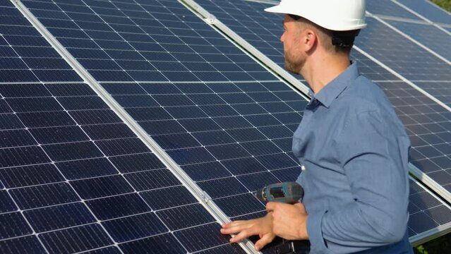Male engineer in protective helmet installing solar photovoltaic panel system using screwdriver. Electrician mounting blue solar module on roof of modern house. Alternative energy ecological concept