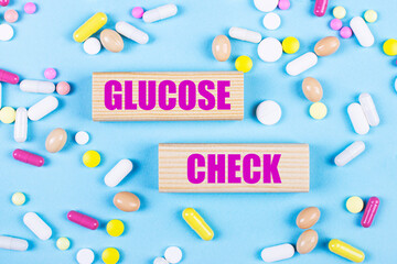 On a light blue background, multi-colored pills and wooden blocks with the text GLUCOSE CHECK. Pharmaceutics. Medical concept.