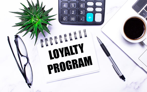 A laptop, glasses, a pen, a calculator, a cup of coffee, a green plant and a white notepad with the text LOYALTY PROGRAM lie on a table with task lighting. View from above. Business concept.