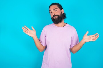 Careless attractive young bearded man wearing violet T-shirt over blue studio background shrugging shoulders, oops.