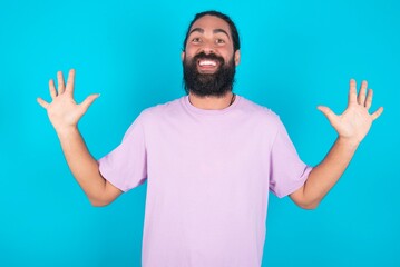 Delighted positive young bearded man wearing violet T-shirt over blue studio background opens mouth  and arms palms up after having great result