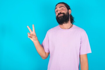 young bearded man wearing violet T-shirt over blue studio background makes peace gesture keeps lips folded shows v sign. Body language concept