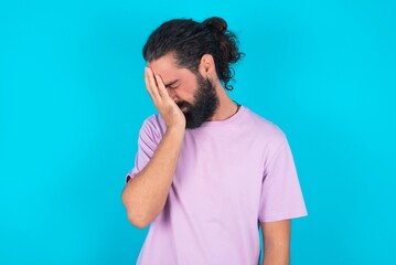 young bearded man wearing violet T-shirt over blue studio background with sad expression covering face with hands while crying. Depression concept.