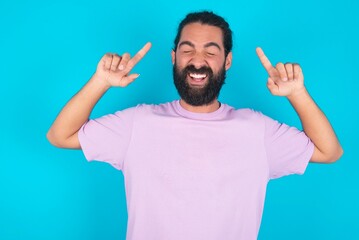 Photo of crazy young bearded man wearing violet T-shirt over blue studio background screaming and pointing with fingers at hair closed eyes
