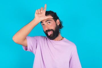 Funny young bearded man wearing violet T-shirt over blue studio background makes loser gesture mocking at someone sticks out tongue making grimace face.