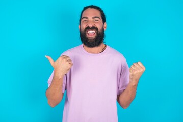 Hooray cool young bearded man wearing violet T-shirt over blue studio background point back empty space hand fist