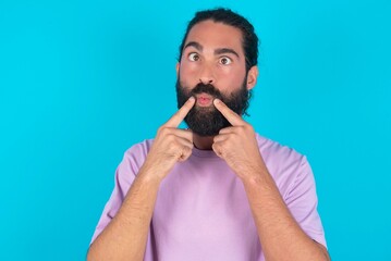 young bearded man wearing violet T-shirt over blue studio background crosses eyes and makes fish lips funny grimace