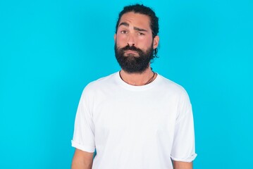 Portrait of dissatisfied young bearded man wearing white T-shirt over blue studio background smirks...