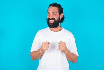 young bearded man wearing white T-shirt over blue studio background clenches fists and awaits for...