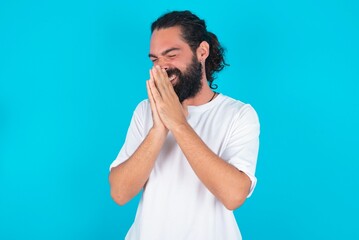 Overjoyed young bearded man wearing white T-shirt over blue studio background laughs joyfully and keeps palms pressed together hears something funny