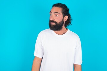 young bearded man wearing white T-shirt over blue studio background stares aside with wondered expression has speechless expression. Embarrassed model looks in surprise