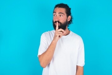 young bearded man wearing white T-shirt over blue studio background makes silence gesture, keeps index finger to lips makes hush sign. Asks not to share secret.