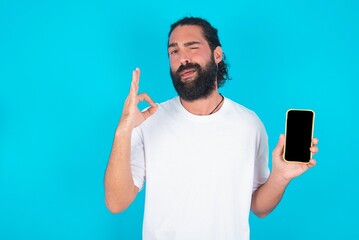 Excited young bearded man wearing white T-shirt over blue studio background showing smartphone blank screen, blinking eye and doing ok sign with hand.  Advertisement concept.