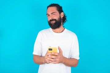 Pleased young bearded man wearing white T-shirt over blue studio background using self phone and looking and winking at the camera. Flirt and coquettish concept.