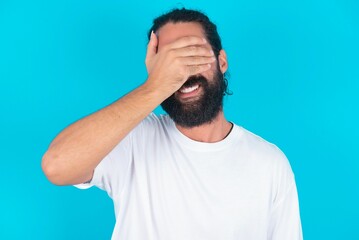 Happy young bearded man wearing white T-shirt over blue studio background closing eyes with hand going to see surprise prepared by friend standing and smiling in anticipation for something wonderful.