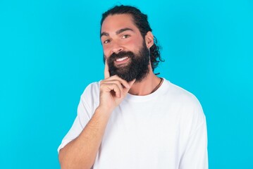 Carefree successful young bearded man wearing white T-shirt over blue studio background touching jawline gazing camera tilting head grinning white teeth delighted. Dental care concept.